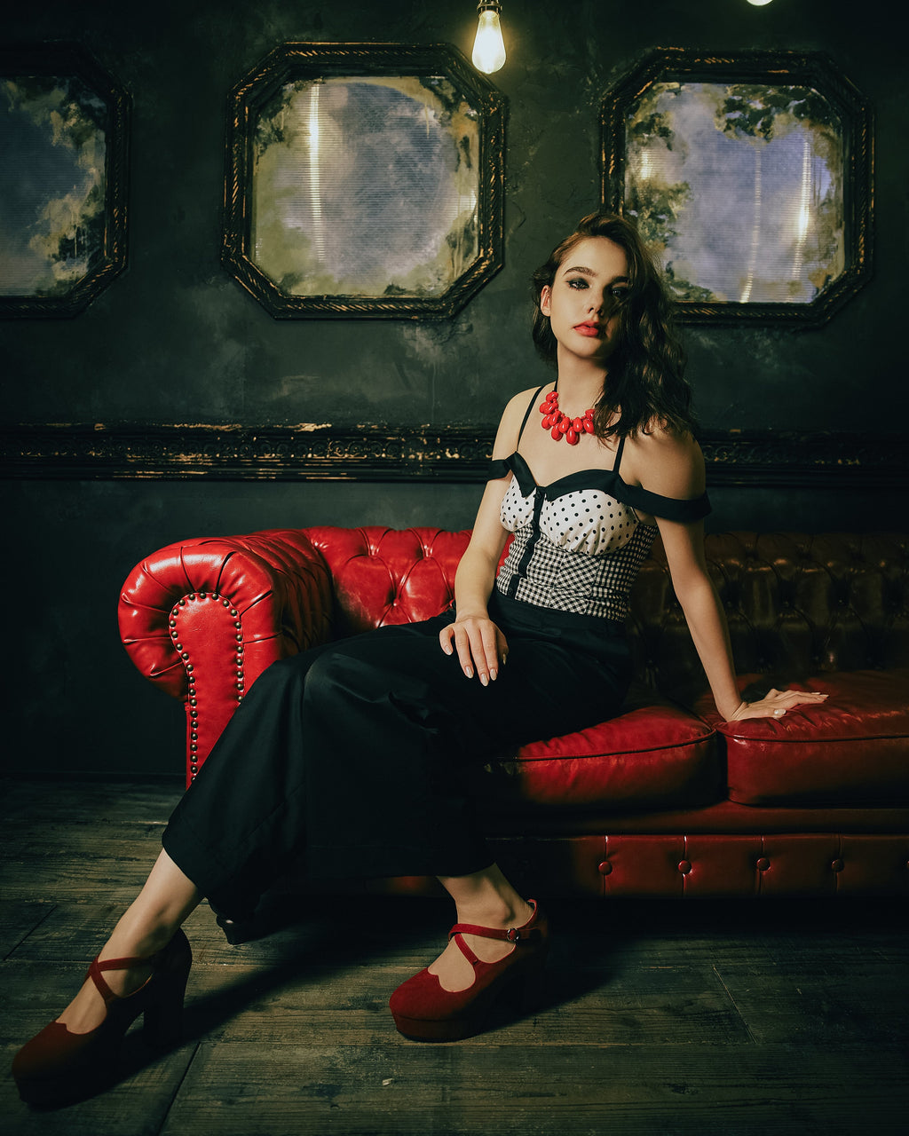 Model wearing the Amy Jumpsuit, polka dot and gingham check with off-shoulder collar. Posing on a red sofa — Atë