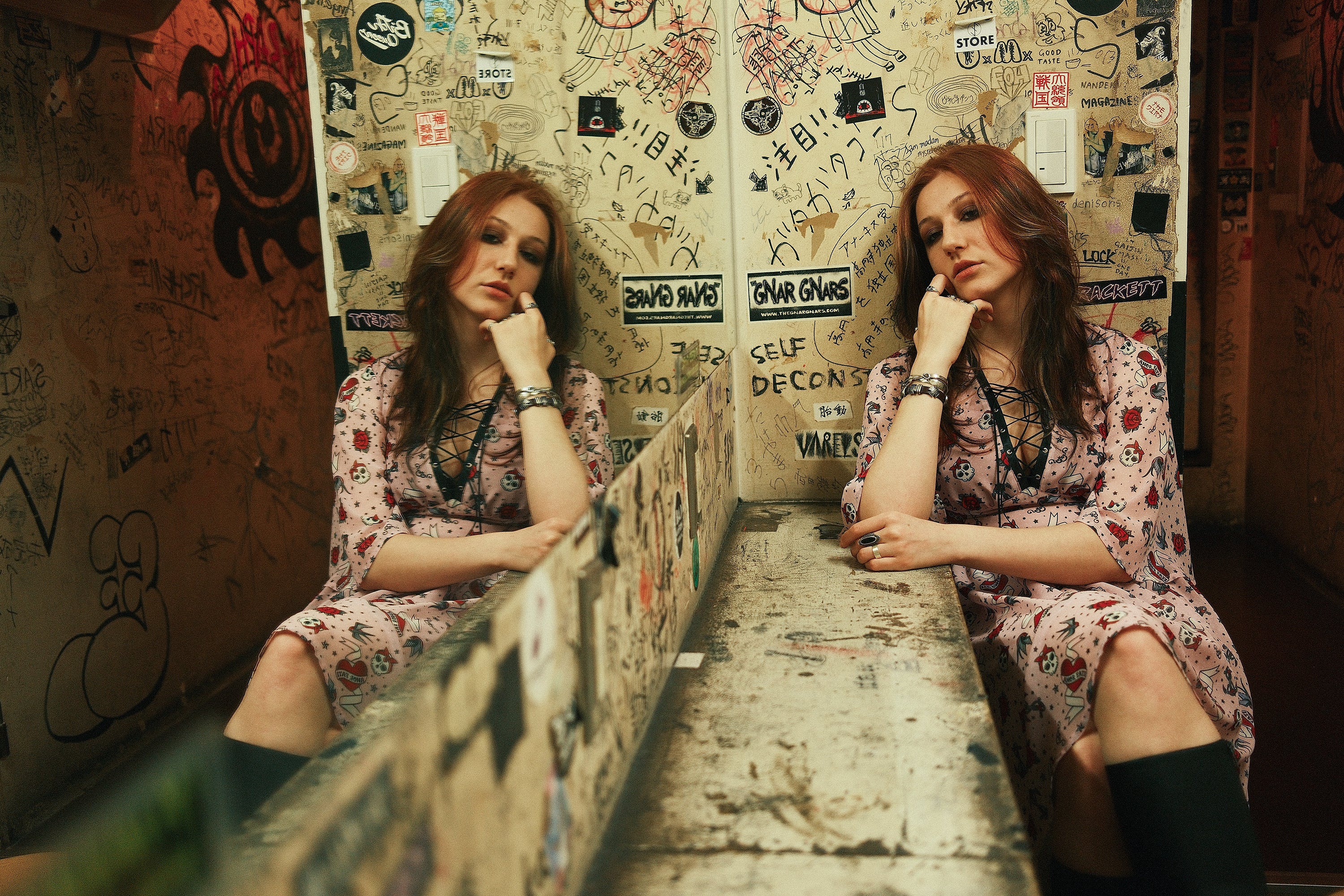Pale pink skull-print chiffon dress with a black lace-up front and flare sleeves, worn seated against a graffiti-covered wall and doubled in a mirror reflection.